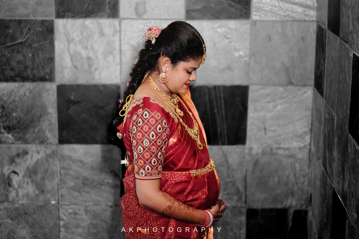 A pregnant woman in a red sari poses in front of a wall during a Baby Shower Photoshoot by AK Photography.
