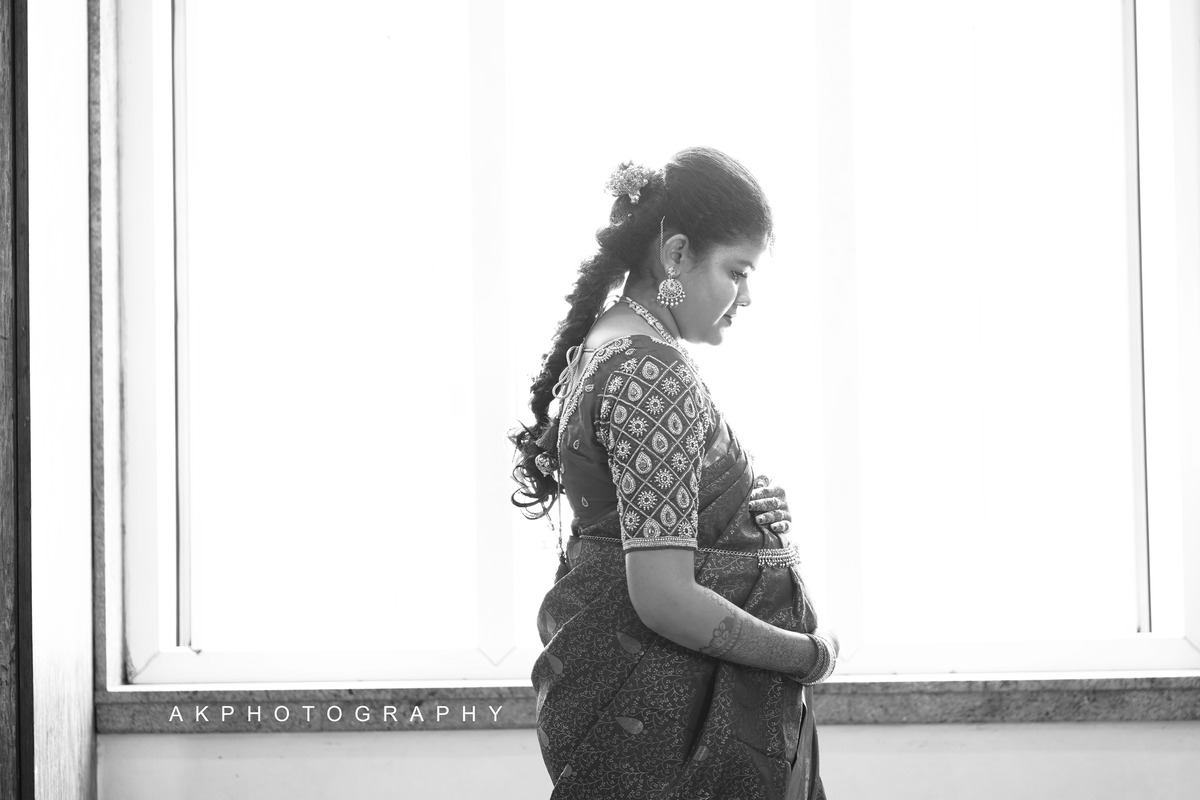 Indian bride in a sari, radiating elegance, stands by the window during a Baby Shower Photoshoot by AK Photography.